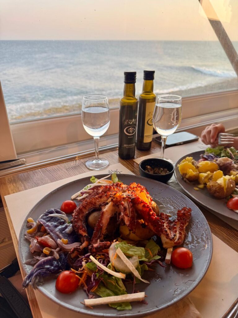 Zwei Teller mit einem Fischgericht und Ausblick aufs Meer von durch die Fenster eines Restaurants in Peniche.